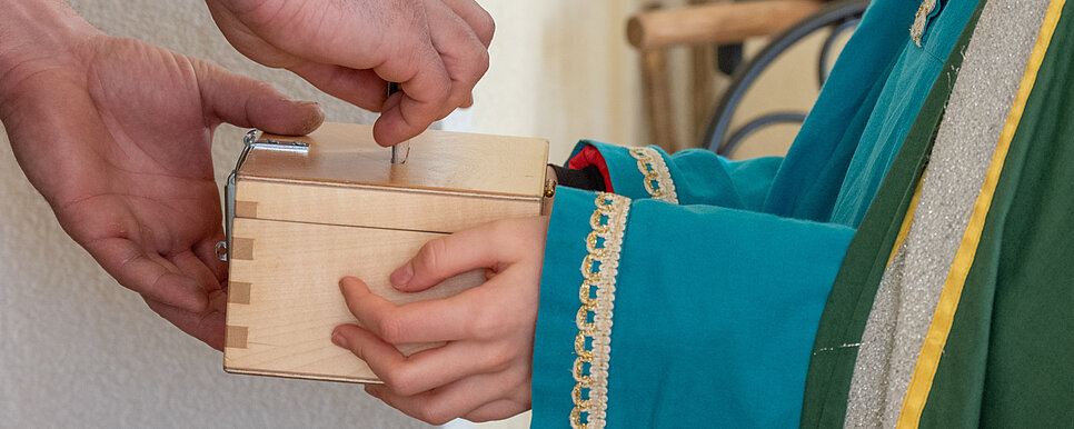 Das Bild zeigt eine Szene, in der eine Person Geld in eine kleine Holzkiste legt, die von einem Kind gehalten wird.