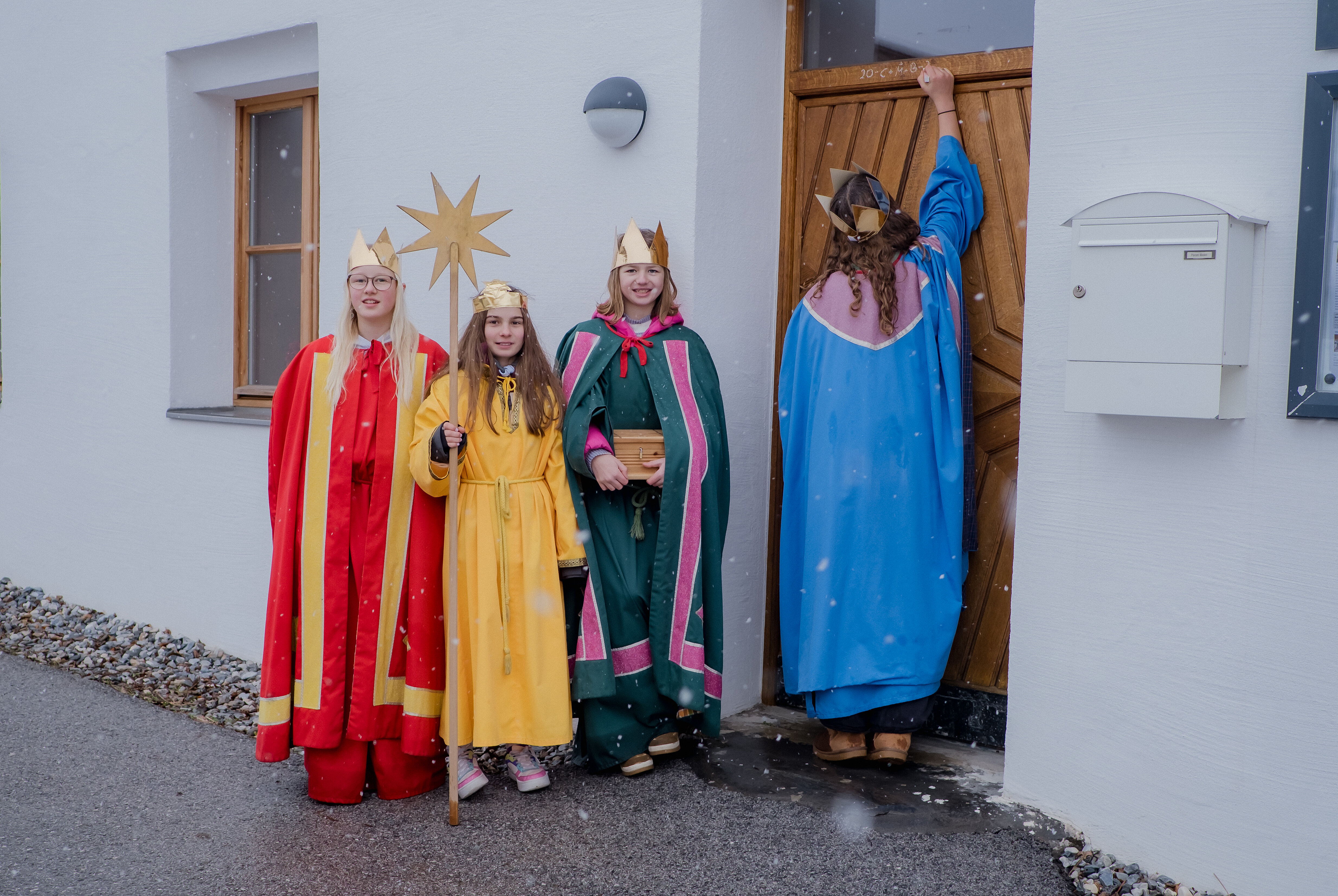 Auf dem Bild sind vier Kinder, in der Kleidung der Sternsinger, zu sehen. Drei von ihnen stehen vor einem weißen Haus und schauen in die Kamera. Sie tragen bunte Gewänder in Rot, Gelb und Grün und jeweils eine Krone auf dem Kopf. Das Kind in Gelb hält einen großen goldenen Stern. Ein viertes Kind, gekleidet in Blau, steht mit dem Rücken zur Kamera und klopft an die Haustür.