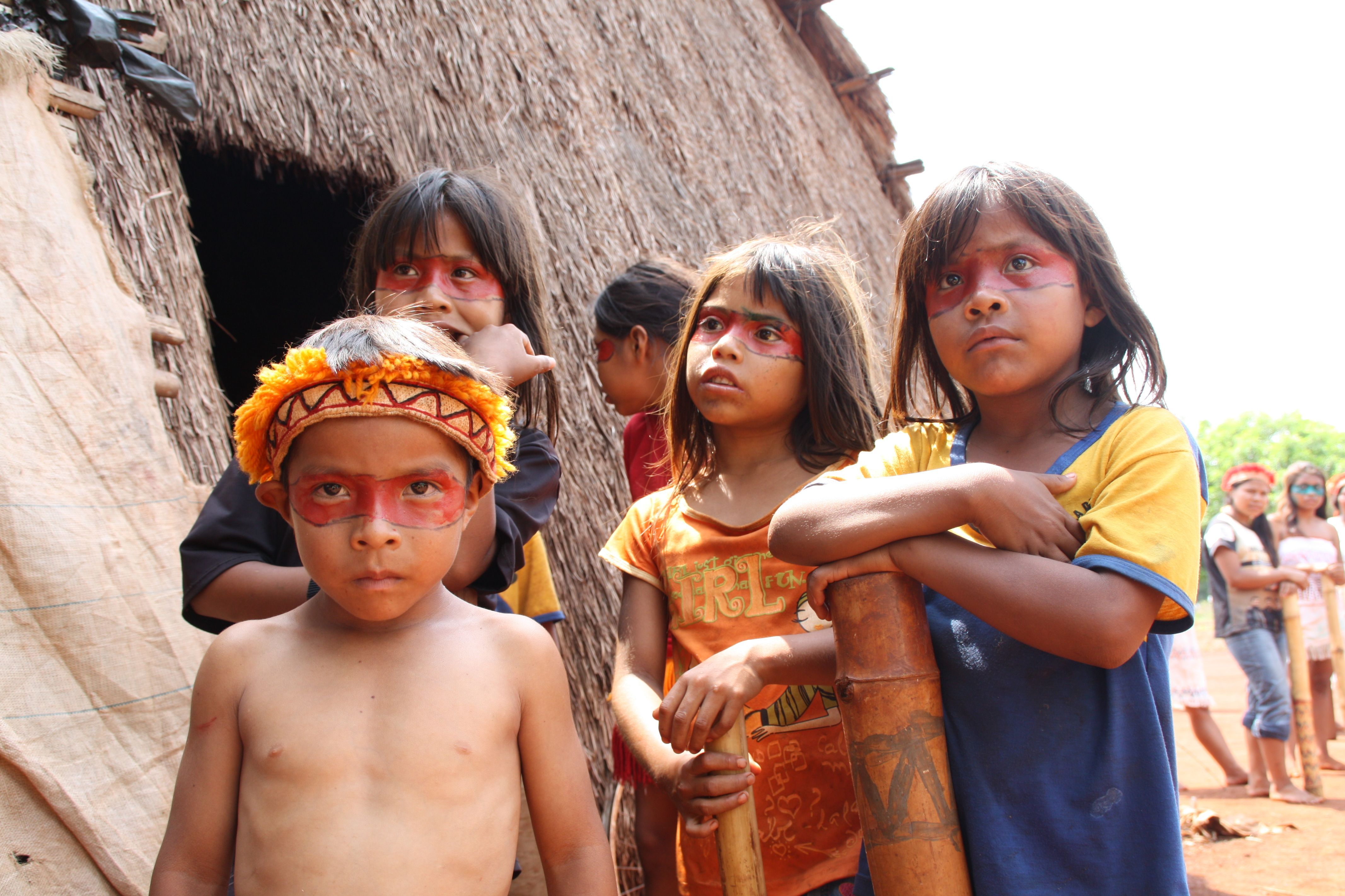 Vier indigene Kinder aus Brasilien stehen draußen vor einer Hütte. Die Augen sind mit einem Band aus roter Farbe umzogen.